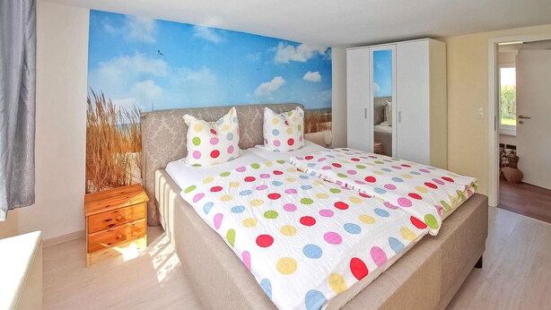 Quarto com uma cama grande, uma mesa de cabeceira de madeira, um guarda-roupa e um mural de paisagem na parede.