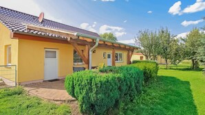 Um edifício amarelo com telhado de telhas, varanda de madeira e um gramado bem cuidado com um cercado aparado.