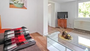 Uma sala de estar com sofá, mesa de centro de vidro, quadro na parede, televisão e janela com vista para árvores.