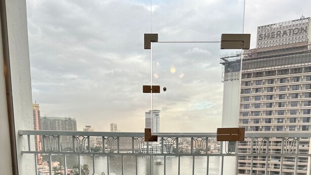 A balcony with two green cushioned chairs, a view of a river with boats, and a cityscape with buildings in the background. - Luxury Flat Stunning Nile View (Giza)
