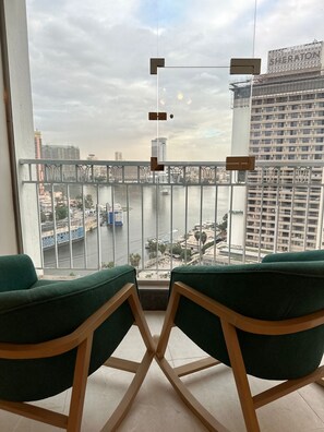A balcony with two green cushioned chairs, a view of a river with boats, and a cityscape with buildings in the background. - Luxury Flat Stunning Nile View (Giza)