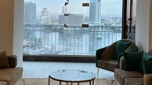 A spacious living room with a large window offering a city view, a modern chandelier, and a round glass coffee table. - Luxury Flat Stunning Nile View (Giza)