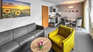 A modern living room with a grey sofa, a yellow armchair, and a small wooden table with a flower. There is a kitchen in the background with a dining area. - Ferienwohnung Trassenheide (Trassenheide (Ostseeba)