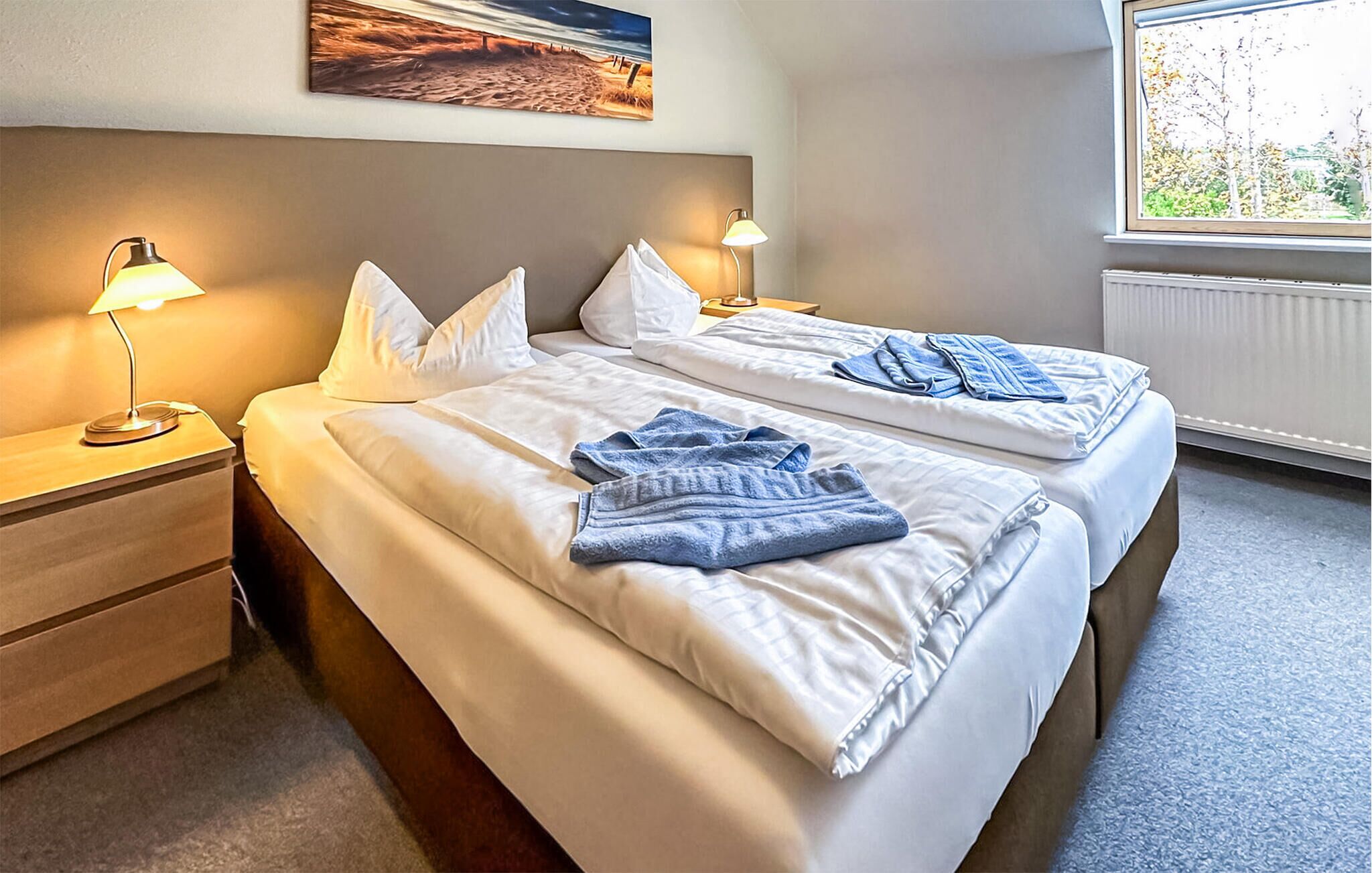 A hotel room with two beds, each with white bedding and blue towels. There are bedside lamps, a wooden nightstand, and a painting on the wall.