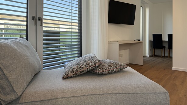 A modern living room with a sofa, patterned pillows, a flat-screen TV mounted on the wall, and a dining area with chairs. - 35qm Serviced Apartment (Töging am Inn)