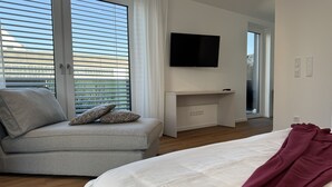 A modern bedroom with a bed, a grey sofa with patterned cushions, a desk with a flat-screen TV, and large windows with blinds. - 35qm Serviced Apartment (Töging am Inn)