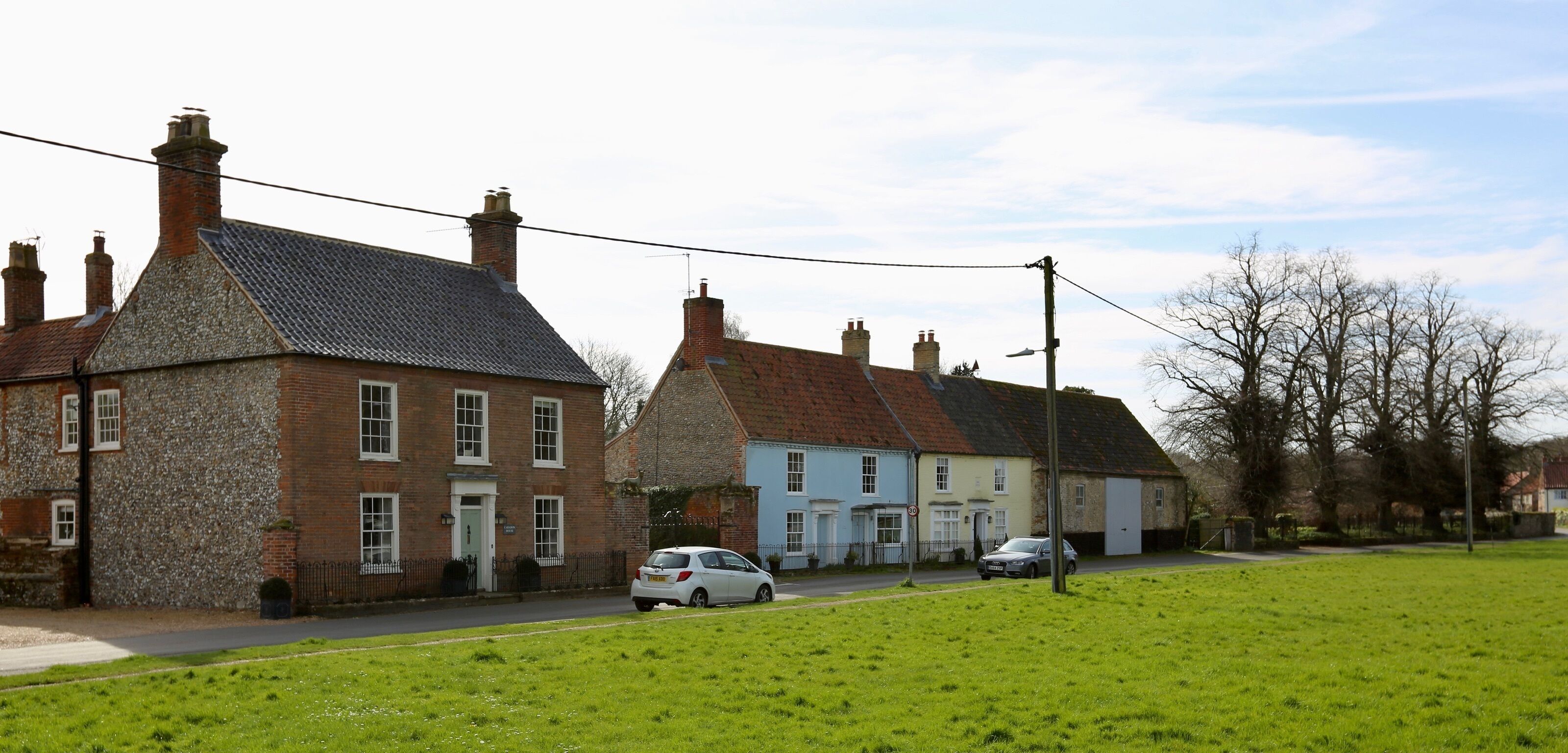 A traditional village street with stone and brick houses, a grassy field, and parked cars.