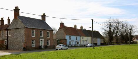 A traditional village street with stone and brick houses, a grassy field, and parked cars.