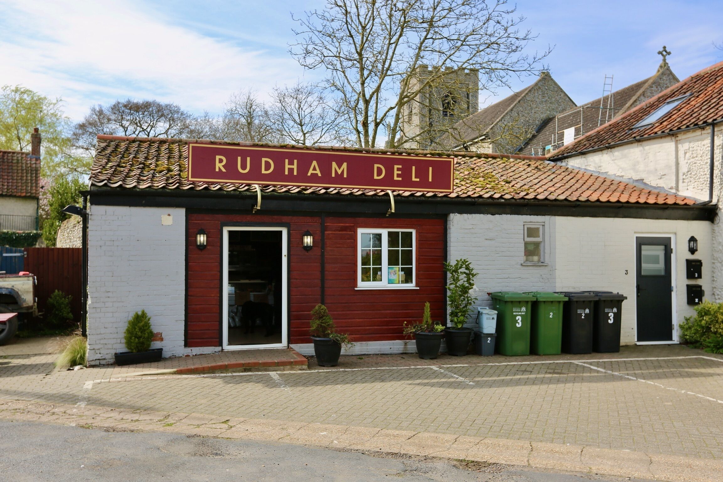 Un bâtiment avec une enseigne indiquant « RUDHAM DELI » et un toit recouvert de tuiles. On peut voir quatre poubelles à l’extérieur, et un petit arbre se trouve près de l’entrée.