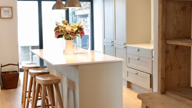 A kitchen with a central island, wooden stools, pendant lights, and a view through large windows.