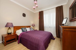 Un dormitorio con una cama de madera, ropa de cama morada, dos lámparas de noche, una mesita de noche, un aparador y un espejo.
