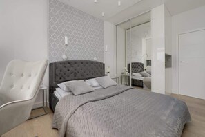 Un dormitorio moderno con un cabecero capitonado gris, una colcha gris acolchada, una silla blanca y un armario con espejo.