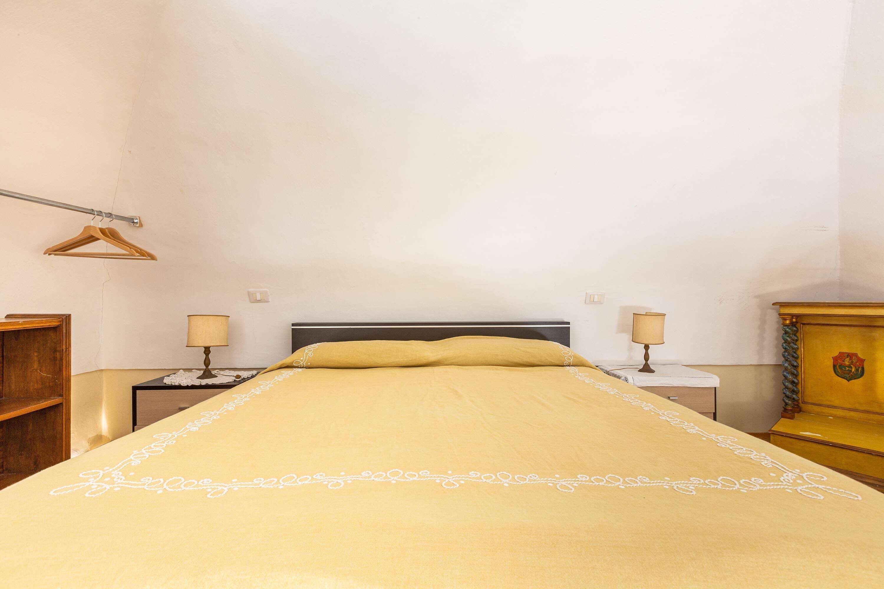 Una cama bien tendida con una colcha amarilla, dos mesitas de noche con lámparas, un aparador de madera y un perchero de madera en la pared.