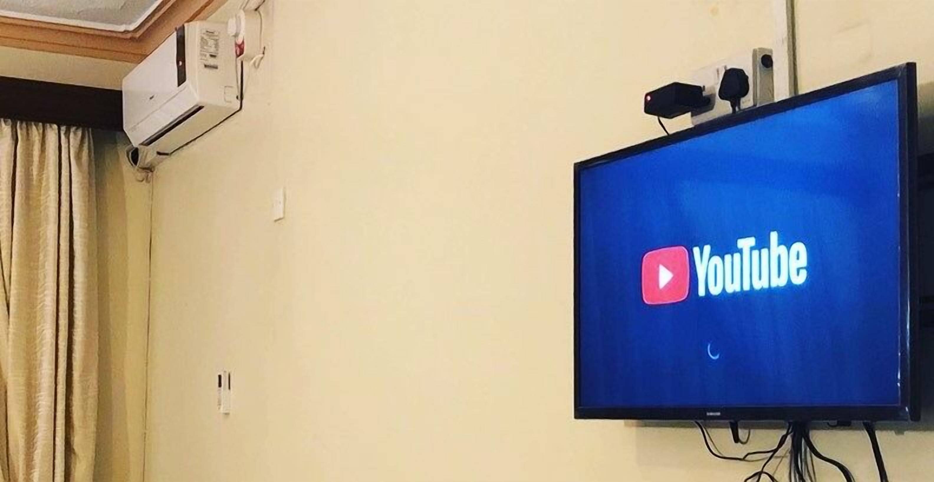 Ein an der Wand befestigter Fernseher zeigt das YouTube-Logo. Eine Decken-Klimaanlage und eine Steckdose sind ebenfalls sichtbar.