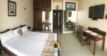 Quarto de hotel com uma cama grande, uma escrivaninha com cadeiras, televisão e porta de acesso a um banheiro.