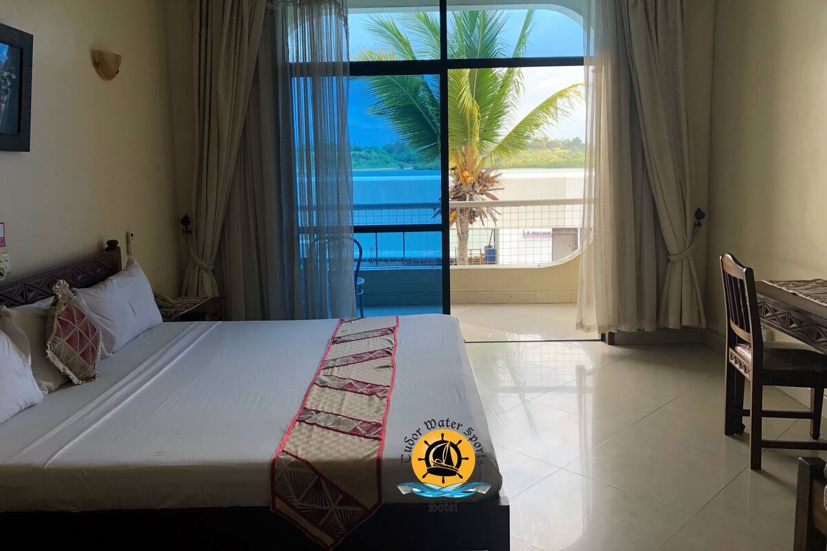 A hotel room with a bed, a desk, and a view of the sea through a large window.