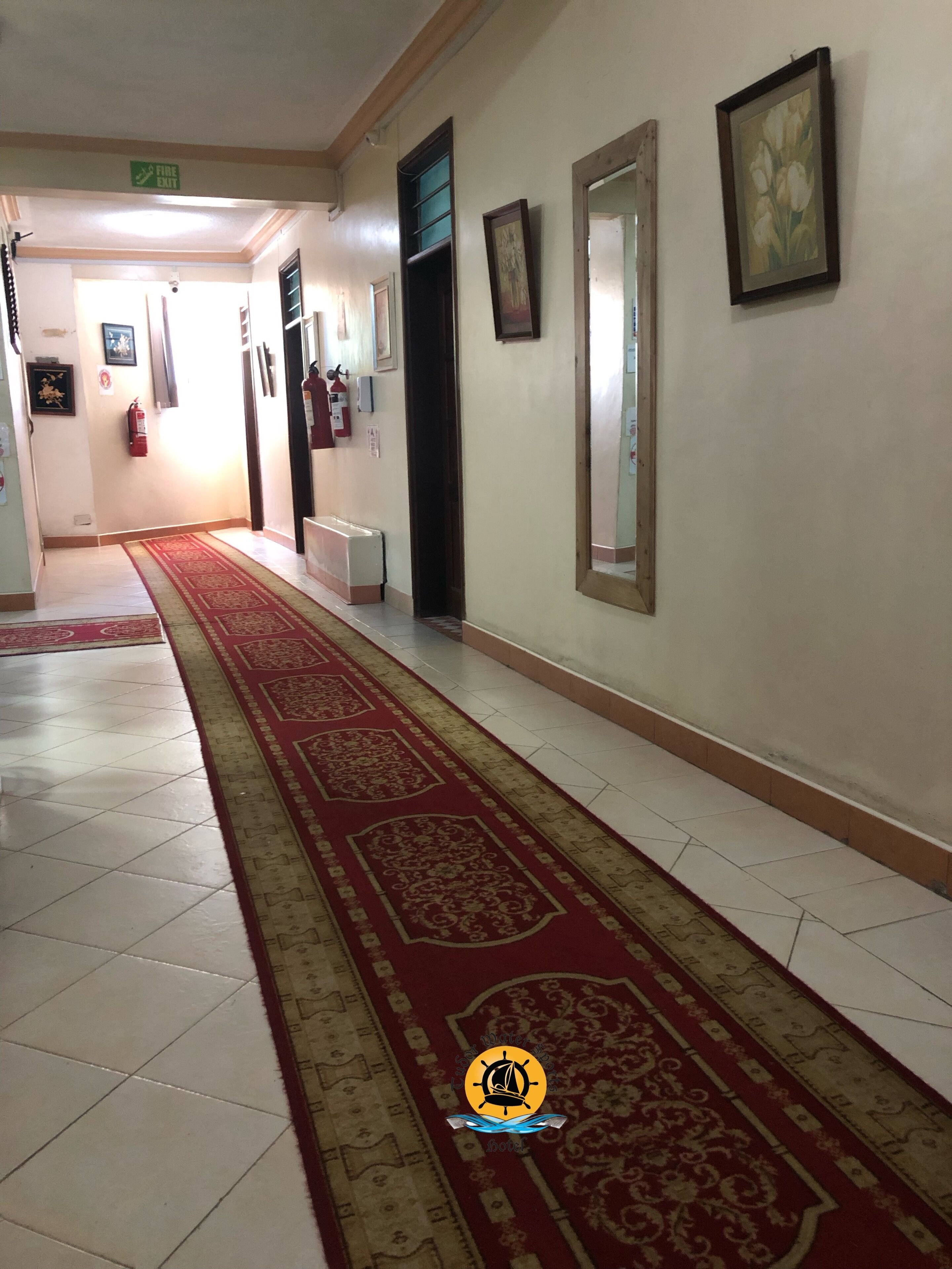 Un pasillo de hotel con alfombra roja, suelo de baldosas, obras de arte enmarcadas en las paredes y un letrero de salida de incendios.