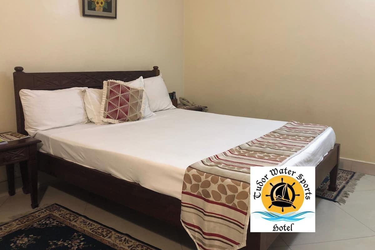 A neatly made bed with a patterned bedspread and pillows, a nightstand with a phone, a framed picture on the wall, and a logo for Tudor Water Sports Hotel.
