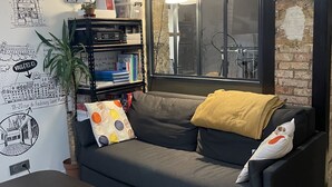 A cozy living room with a grey sofa, a coffee table, and a bookshelf. There's a window with a view of a bicycle and a brick wall.