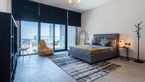 Een moderne slaapkamer met een groot bed, een stoel, een vloerlamp en uitzicht op de stad door grote ramen.