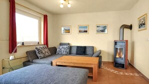 Una sala de estar acogedora con un sofá, un reposapiés y una mesa de madera. Hay una chimenea con su correspondiente chimenea, y tres cuadros enmarcados en la pared.