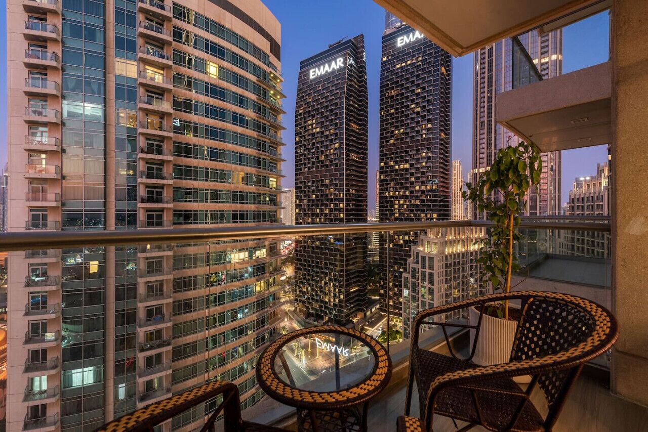 Ein Balkon mit Korbstühlen und einem Blumentopf, mit Blick auf eine Skyline moderner Gebäude und einen klaren Himmel.