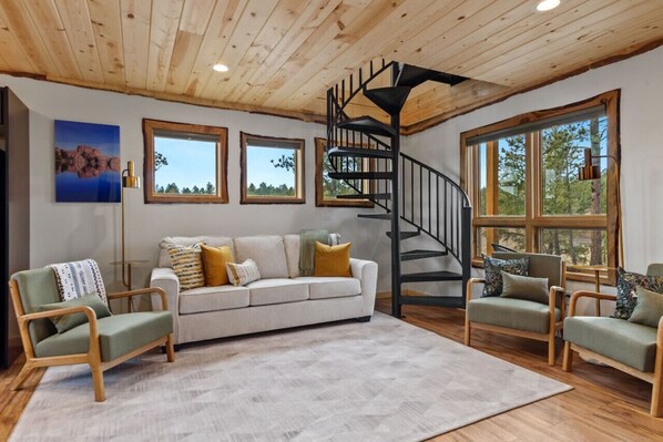 Un salón espacioso con un sofá blanco, dos sillones verdes, una escalera espiral de madera y ventanas amplias con vistas a árboles.