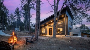 Una casa moderna de dos pisos amb una llar de foc, seients exteriors i vistes a la forest circumdant.