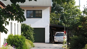 Een moderne, twee verdiepingen tellende woning met een garagepoort, omringd door weelderige beplanting en een oprit naar een geparkeerde auto.