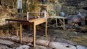 A rustic stone-walled room with a wooden table, a chair, a basket, and a pot on the floor.