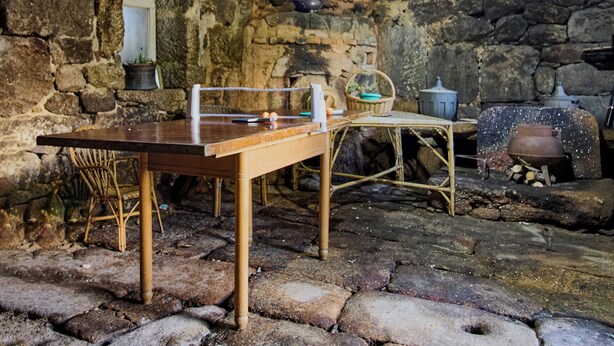 Une pièce rustique aux murs de pierre, avec une table en bois, une chaise, un panier et un pot posé sur le sol.