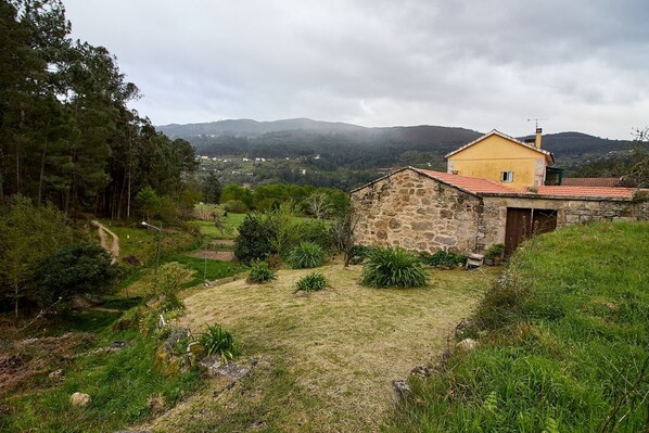 A stone building with a red roof, surrounded by a grassy area and trees, with a mountainous landscape in the background. - THE HOUSE OF MILA (Crecente)