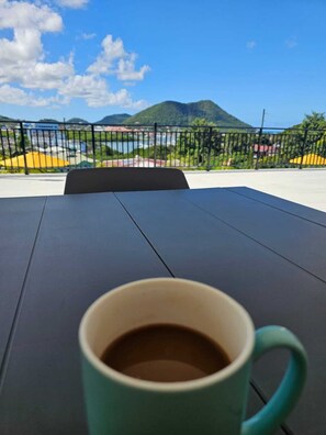 Une table avec une tasse de café, une vue sur une montagne et une zone délimitée par une clôture, avec des bâtiments en arrière-plan.