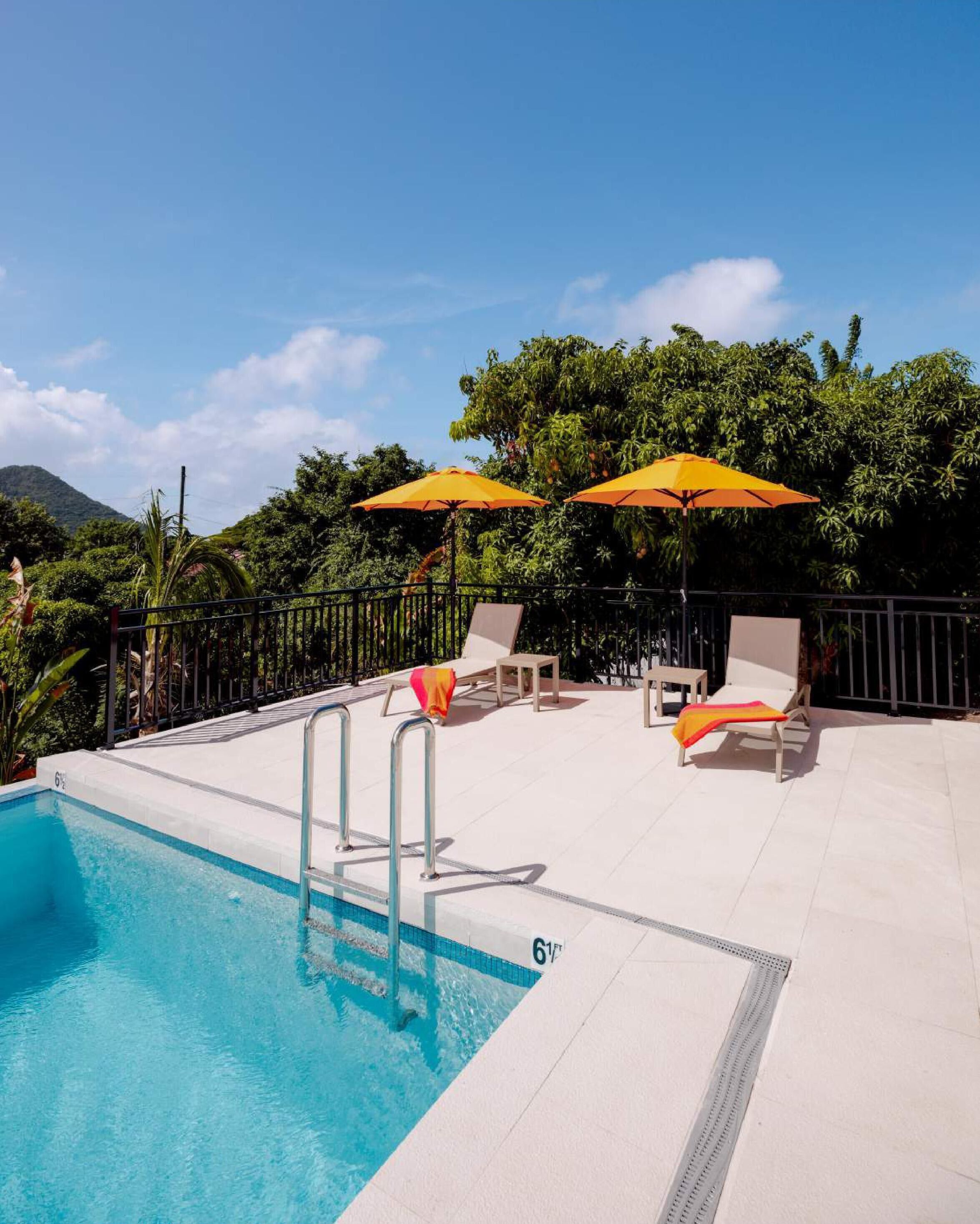 A swimming pool with two lounge chairs and two umbrellas, surrounded by a fence and lush greenery.