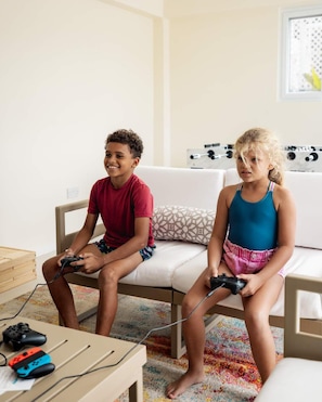 Deux enfants jouent à des jeux vidéo sur un canapé dans un salon, où se trouvent un tapis à motifs et une petite table sur laquelle reposent des manettes de jeu.