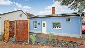 Une maison à deux étages, à l’extérieur bleu, avec une porte blanche et un petit jardin à l’avant, agrémenté d’une clôture en bois et d’un chemin de gravier.