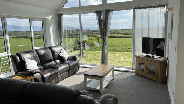 A modern living room with a leather sofa, a wooden coffee table, and a television on a wooden cabinet. Large windows offer a view of a green landscape. - Beautiful, Private Farm Cottage in Anglesey (Pentraeth)