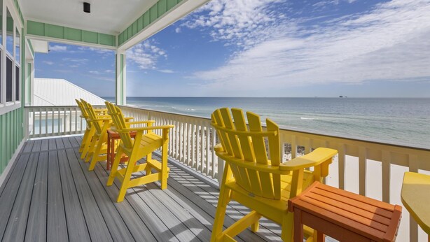 Une terrasse en bois avec des fauteuils Adirondack jaunes et une table, donnant sur une plage, l’océan et un ciel dégagé.