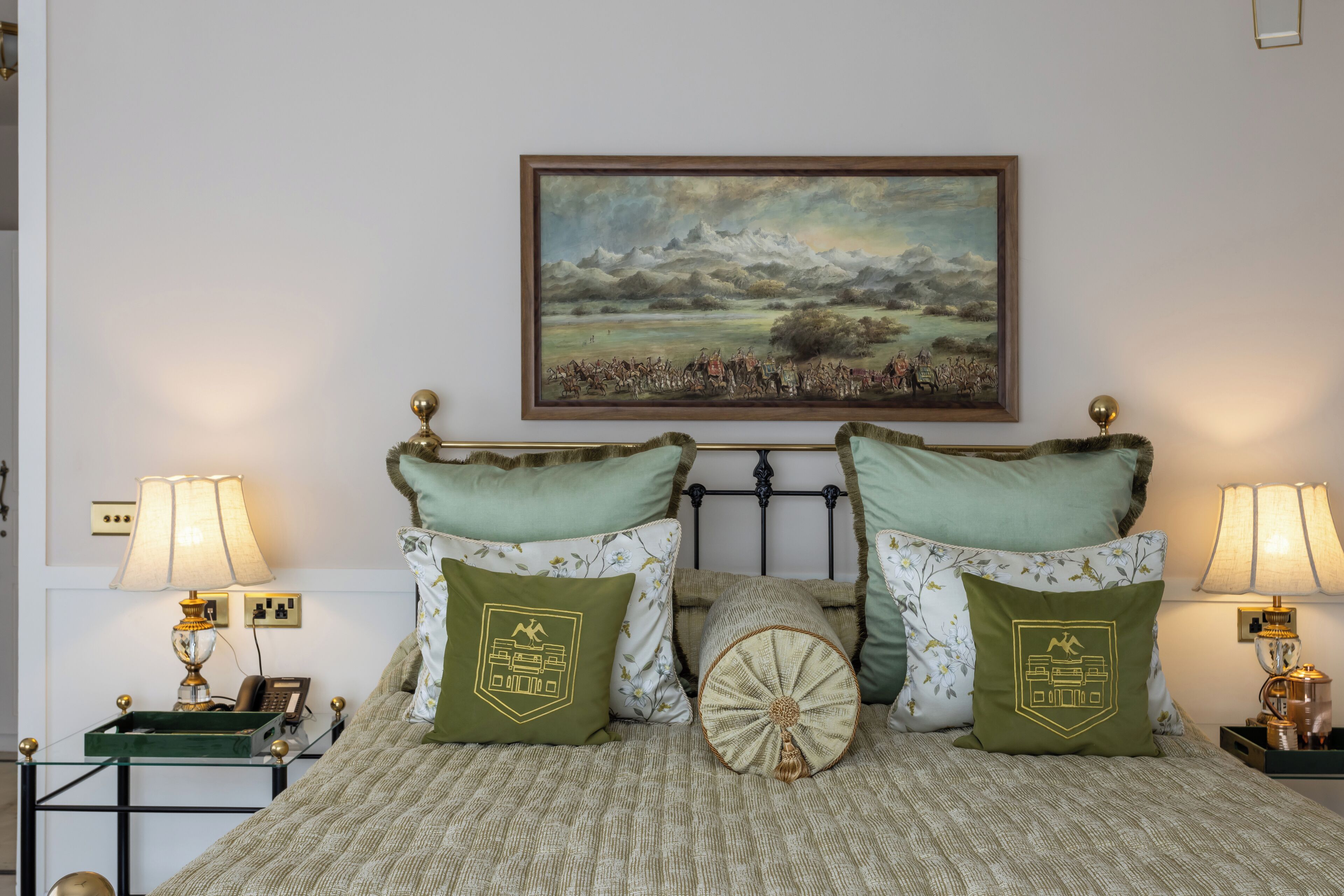 Un dormitorio ordenado con cama, dos almohadas verdes, una almohada decorativa, un cuadro enmarco de un paisaje y lámparas de noche.