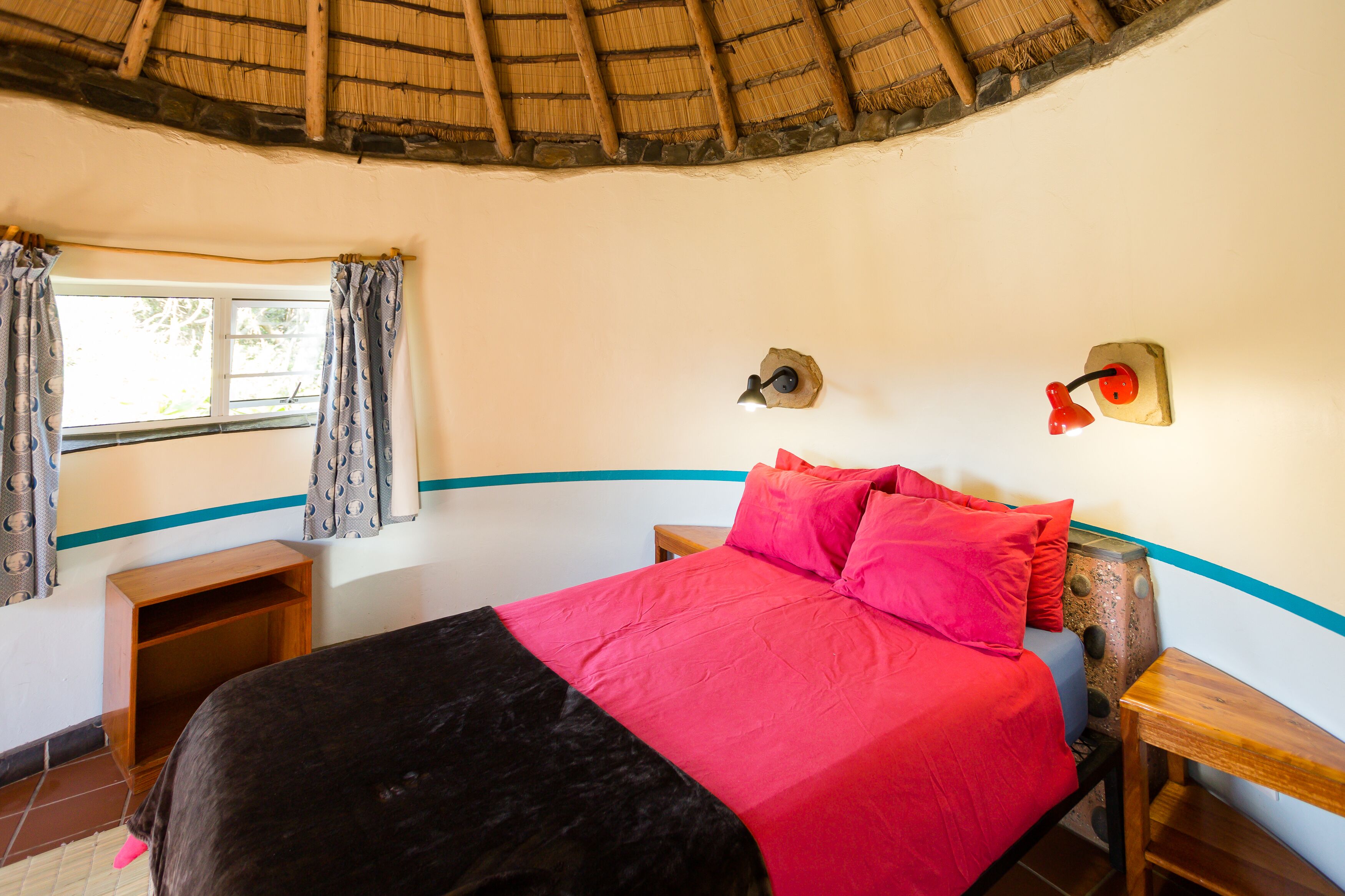 A room with a bed, a window with curtains, a wall-mounted lamp, and a small wooden table.