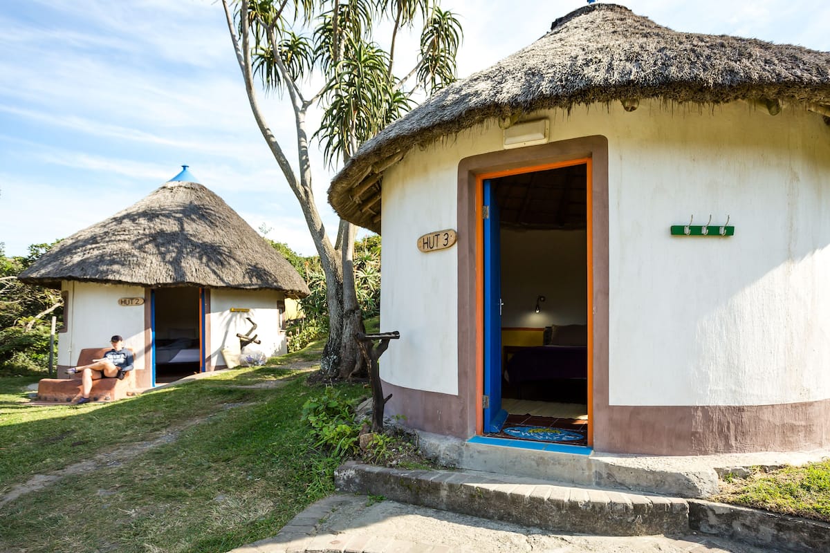 Eine reetgedeckte Hütte mit der Aufschrift 'HUT 3', sie hat eine blaue Tür und eine kleine Veranda.
