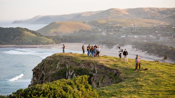 Un groupe de personnes se trouve sur une falaise herbeuse, d’où elles dominent un paysage côtier composé de collines et, au loin, d’une plage.
