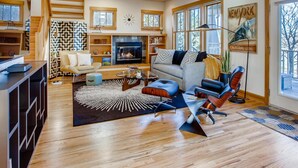 A spacious living room with a fireplace, a sofa, a coffee table, and a large window with a view of trees. - Brilliant Posh Home - Backs to Poudre River Trail (Fort Collins)