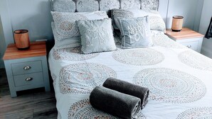 A neatly made bed with patterned bedding, two bedside tables with lamps, and a wooden nightstand with drawers. - Serenity Holiday Let in South Chard (Chard)