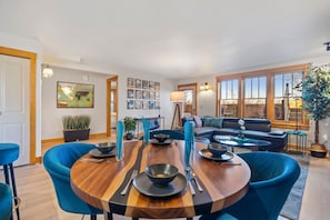 Ein Essbereich mit einem Holztisch, gedeckten Plätzen für vier Personen, blauen Polsterstühlen und einer schwarzen Couch im Hintergrund.