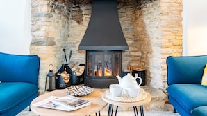 A cozy room with a stone fireplace, two blue sofas, a small round table with a magazine, a kettle, and a tray of logs. - Gable Cottage  - Dog Friendly, Meysey Hampton near Cirencester (Meysey Hampton)