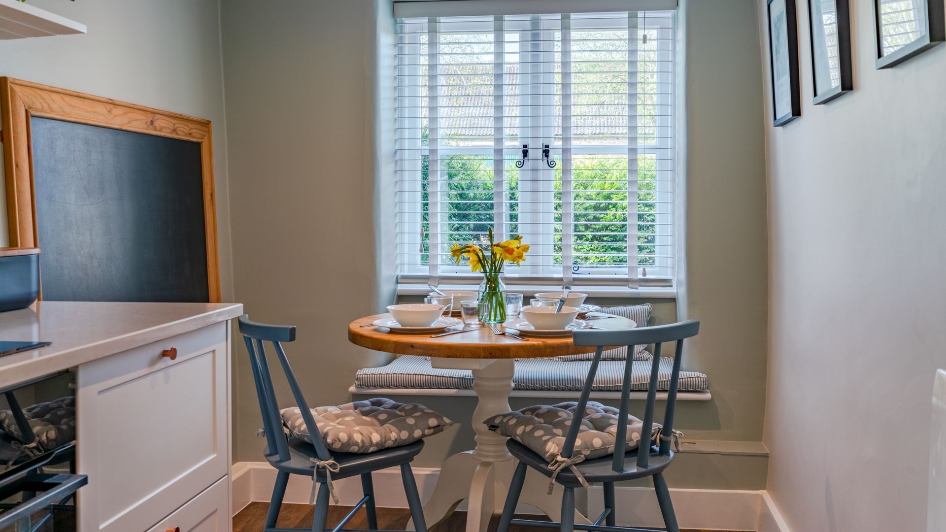 Une petite cuisine avec un tableau noir, une fenêtre avec des stores, une table à manger avec des chaises et un vase rempli de fleurs.