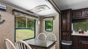 A compact RV kitchen with a dining area, including a table and chairs, a window, and a small trash can. - Holiday Haven has Water Access with Private Boat Launch on Lake Guntersville! (Scottsboro)