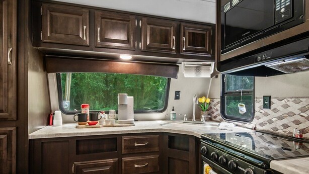 A compact RV kitchen with dark wood cabinets, a stove, and a window with a view of greenery. - Holiday Haven has Water Access with Private Boat Launch on Lake Guntersville! (Scottsboro)