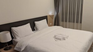 Een keurig opgemaakt bed met witte lakens en een gevouwen handdoek, een bedlampje en een raam met dunne gordijnen.